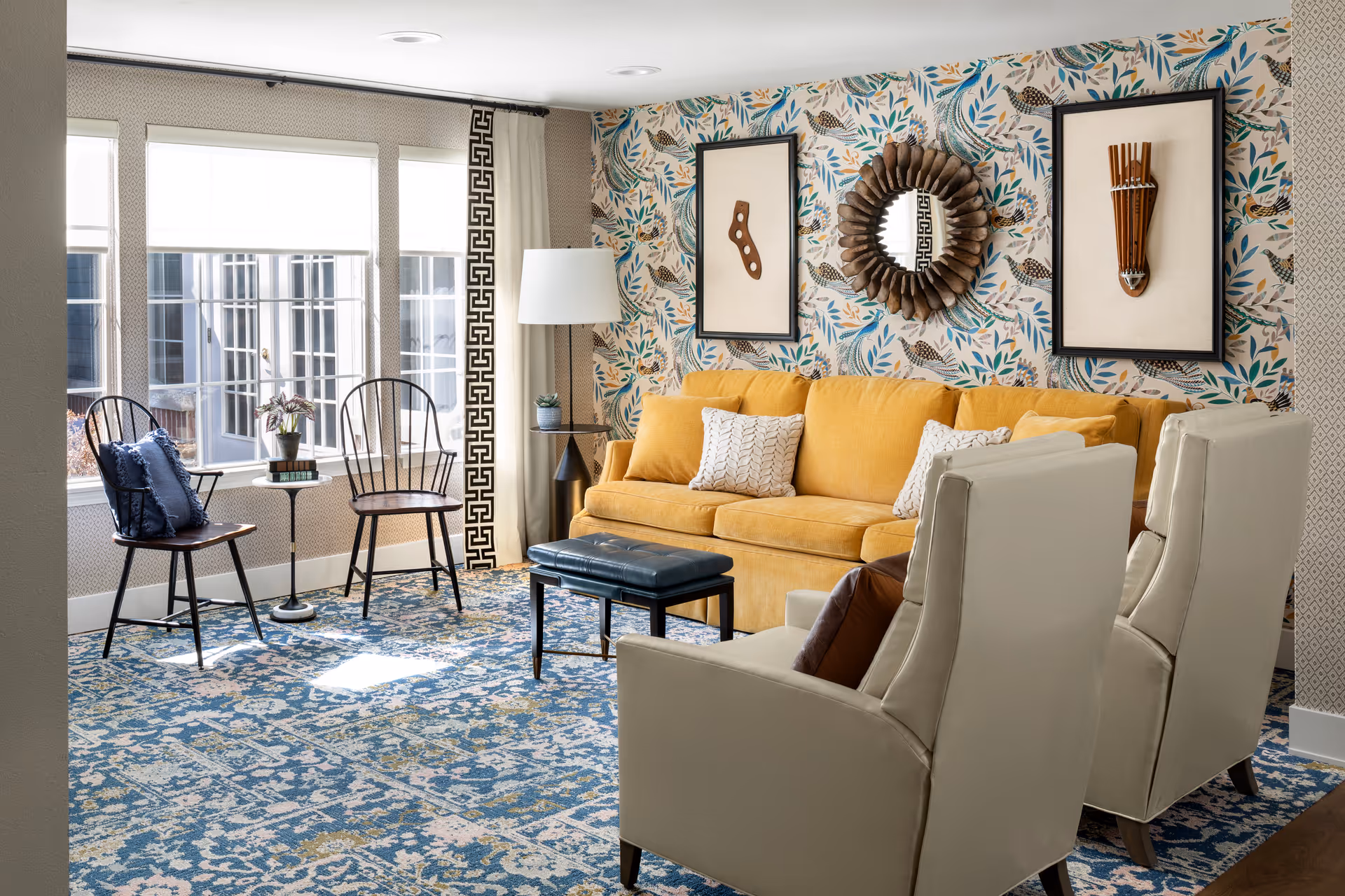 A bright and cozy living room with a mustard yellow sofa adorned with white and patterned pillows, two cream-colored armchairs, and two wooden chairs with cushions near a window. The room features a patterned blue and beige carpet, a floor lamp, a small side table with a plant, and a decorative wall with framed art and a round mirror.