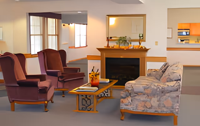 A cozy living room area in Grandview Estates featuring two purple armchairs and a patterned sofa arranged around a wooden coffee table with books and a small plant. A fireplace with a mirror above it is centered on the back wall, and a kitchen area is visible through a pass-through window on the right.