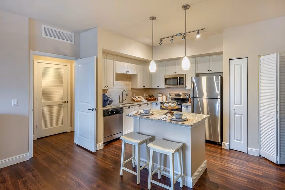 Bright modern kitchen with an island and two stools, stainless steel appliances, white cabinets, and pendant lights.