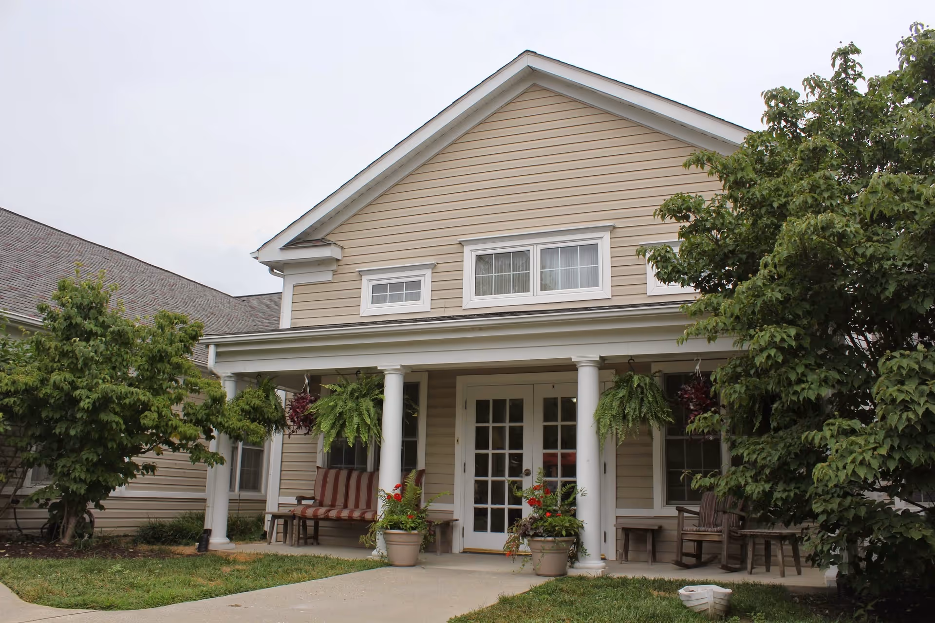 Front exterior view of a beige assisted living facility building with white columns supporting a small porch. There are two potted plants with flowers on either side of the entrance, hanging green ferns, and outdoor seating including a bench and chairs. Trees and shrubs surround the entrance area.