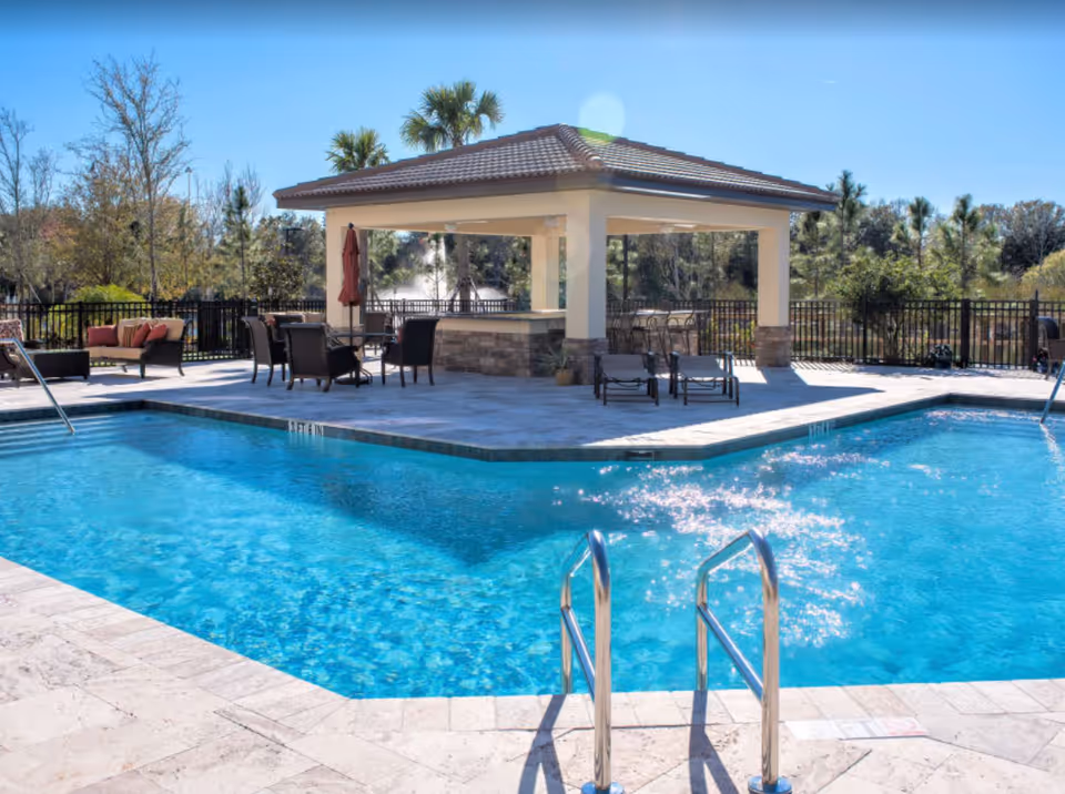 Outdoor swimming pool with clear blue water, surrounded by a stone deck with lounge chairs, tables, and a covered pavilion with seating. Trees and greenery are visible in the background under a clear blue sky.