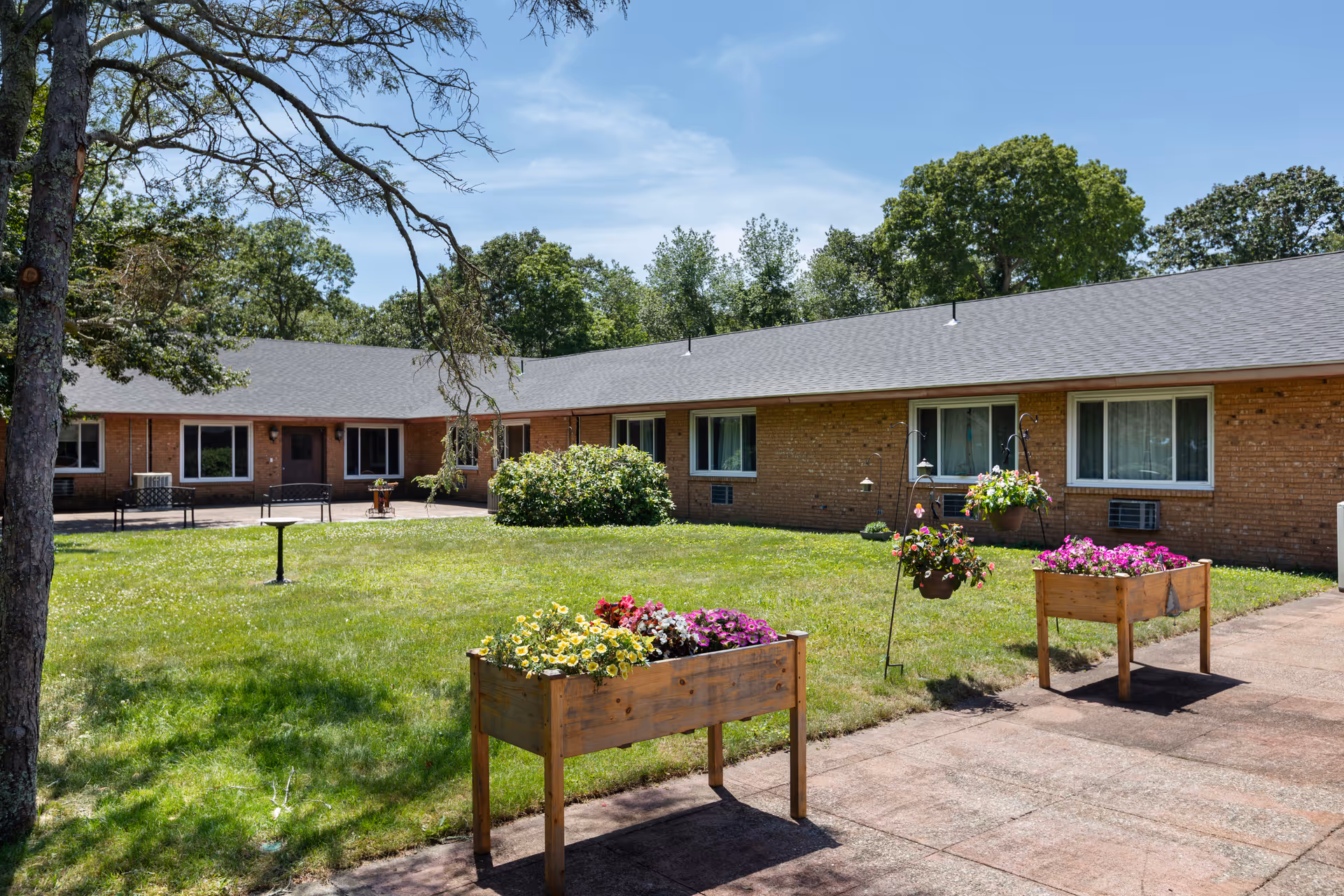 Single-story brick building surrounding a grassy courtyard with raised flower planters, hanging baskets, and patio seating under a clear sky.