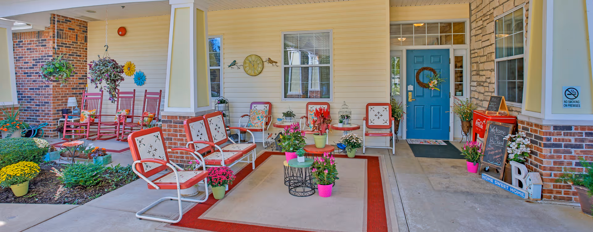 Covered outdoor porch area with red and white metal chairs and benches arranged around small tables with potted plants. Hanging plants and red rocking chairs are visible on the left side. The porch has a blue door with a wreath, a welcome chalkboard sign, and various decorative flowers and plants. The walls are a combination of brick and light yellow siding.