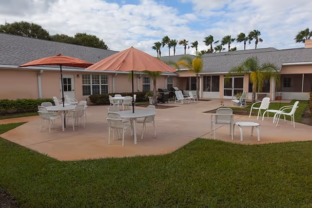 Outdoor patio area with several round tables and chairs, some tables shaded by large umbrellas. The patio is surrounded by a single-story building with windows and doors, and there are palm trees and green grass around the patio under a partly cloudy sky.