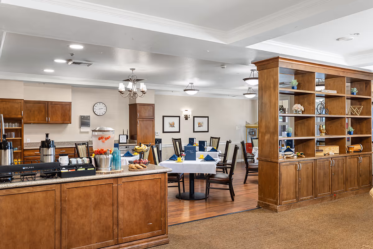 A dining area in a senior living facility featuring wooden cabinets and a counter with beverage dispensers and snacks. Several tables are set with white tablecloths, blue napkins, and menus, surrounded by wooden chairs. The room has warm lighting with chandeliers and wall sconces, and a large wooden shelving unit partially divides the space.