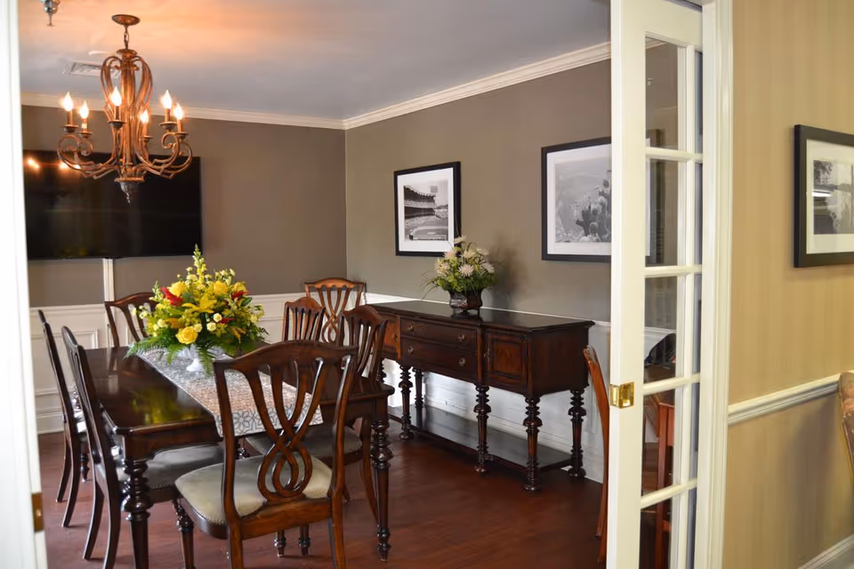 A formal dining room with a long wooden table and chairs, chandelier overhead, a floral centerpiece, and a wooden sideboard against the wall.