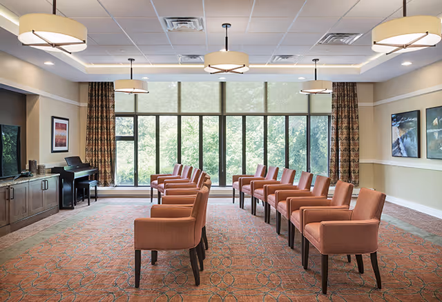 A spacious, well-lit room with large floor-to-ceiling windows covered partially by roller shades. The room features two rows of orange upholstered chairs facing a large flat-screen TV mounted on a wooden cabinet. There is a piano in the corner and framed artwork on the walls. The ceiling has multiple modern circular light fixtures.