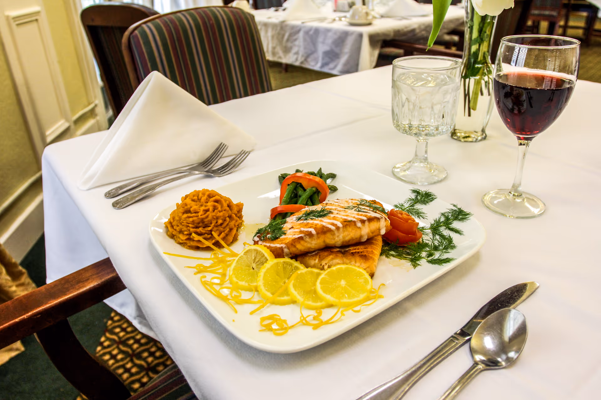 Plated salmon entrée with lemon slices, mashed sweet potato, vegetables, a glass of red wine and water on a white-tablecloth dining table in a dining room.
