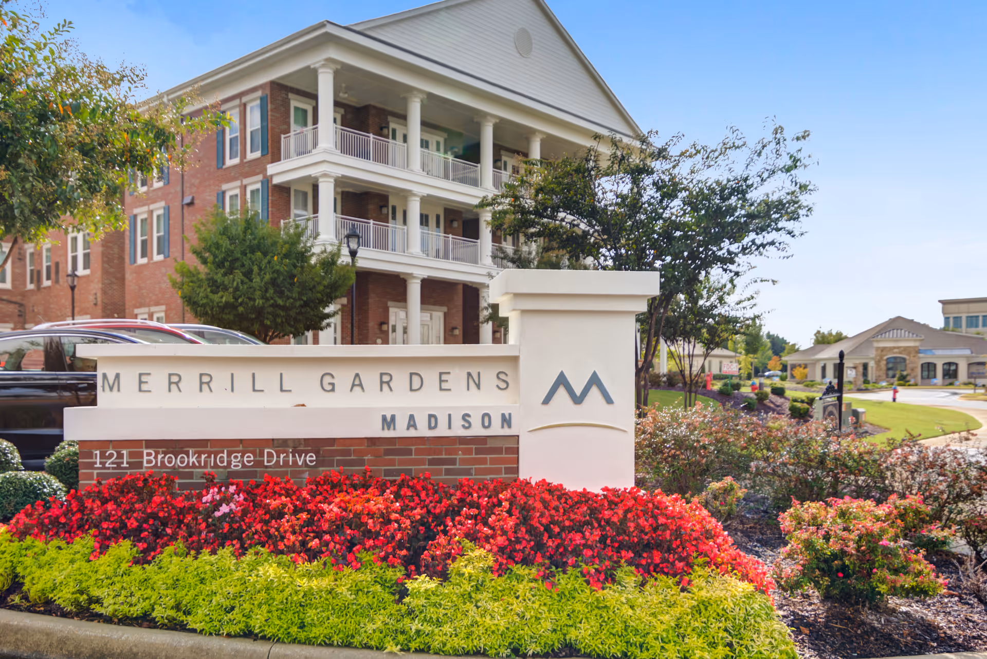 Entrance sign reading "Merrill Gardens Madison" with colorful landscaped flowerbeds and the residential building in the background.
