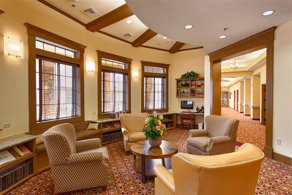 A cozy sitting area in a senior living facility with four armchairs arranged around a round wooden coffee table with a flower arrangement. The room features large windows with wooden blinds, built-in bookshelves filled with books, and a small desk with a computer. The carpet has a floral pattern, and the walls are painted in warm tones with wooden trim and ceiling beams.