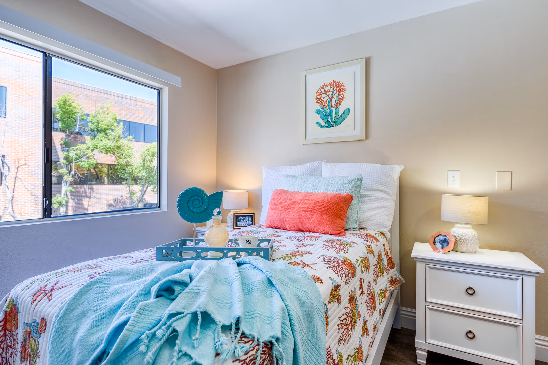Bright single bedroom with a made bed, colorful pillows and throw, a nightstand with lamp, and a window showing trees and a brick building outside.