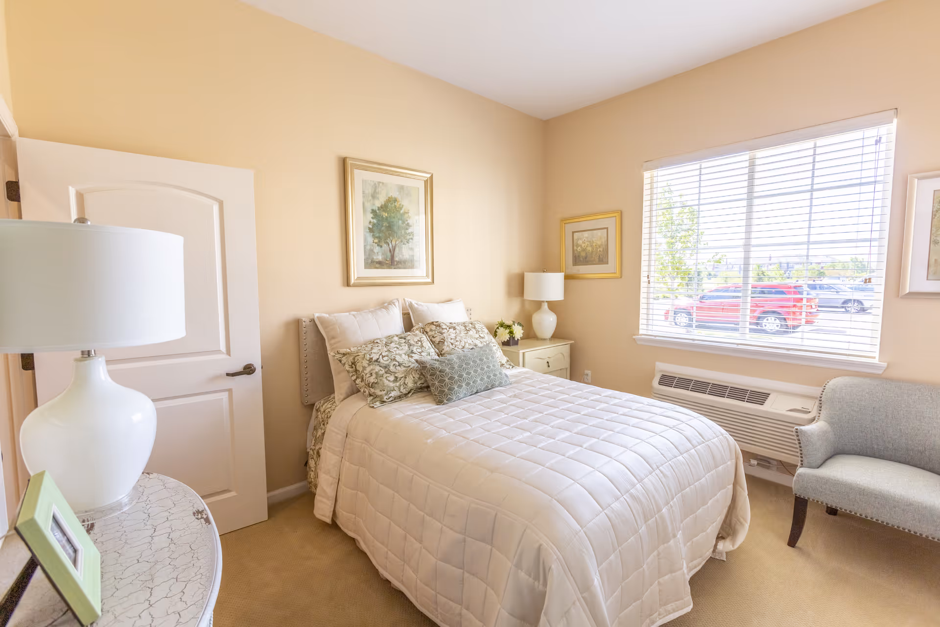 A bright and cozy bedroom with a neatly made bed featuring a white quilt and decorative pillows. The room has beige walls, a window with blinds letting in natural light, a small armchair, two bedside tables with lamps, and framed artwork on the walls.