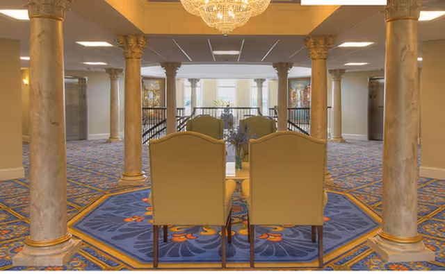 Interior view of a senior living facility lobby area with ornate columns, a chandelier, and a patterned blue carpet. Four upholstered chairs are arranged around a small table in the center, with large windows and artwork visible in the background.