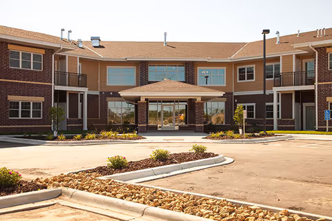 Front entrance of a two-story brick and siding senior apartment building with a covered porte-cochere and landscaped driveway.