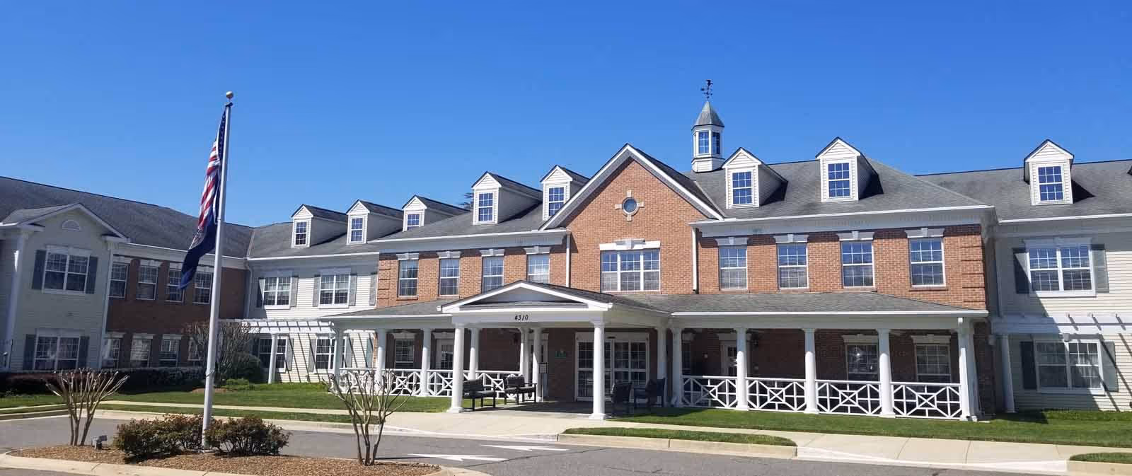 Front exterior of a large brick-and-siding senior living facility with a covered entrance, columns, porch seating, and a flagpole under a clear blue sky.