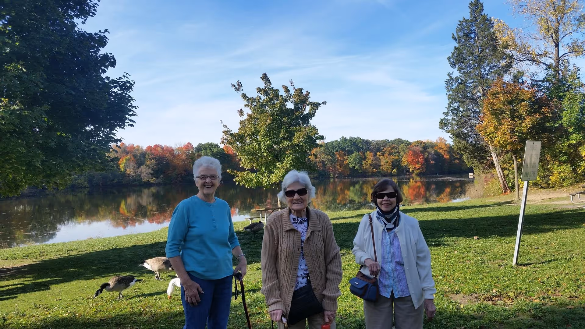 Three elderly women standing on a grassy area near a calm lake with trees showing autumn colors in the background. Two of the women are wearing sunglasses, and one is holding a walking cane. There are geese grazing on the grass nearby under a clear blue sky.