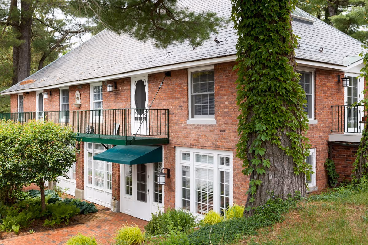 Brick two-story building with a green awning, upper balcony, large ivy-covered tree, and surrounding landscaping.
