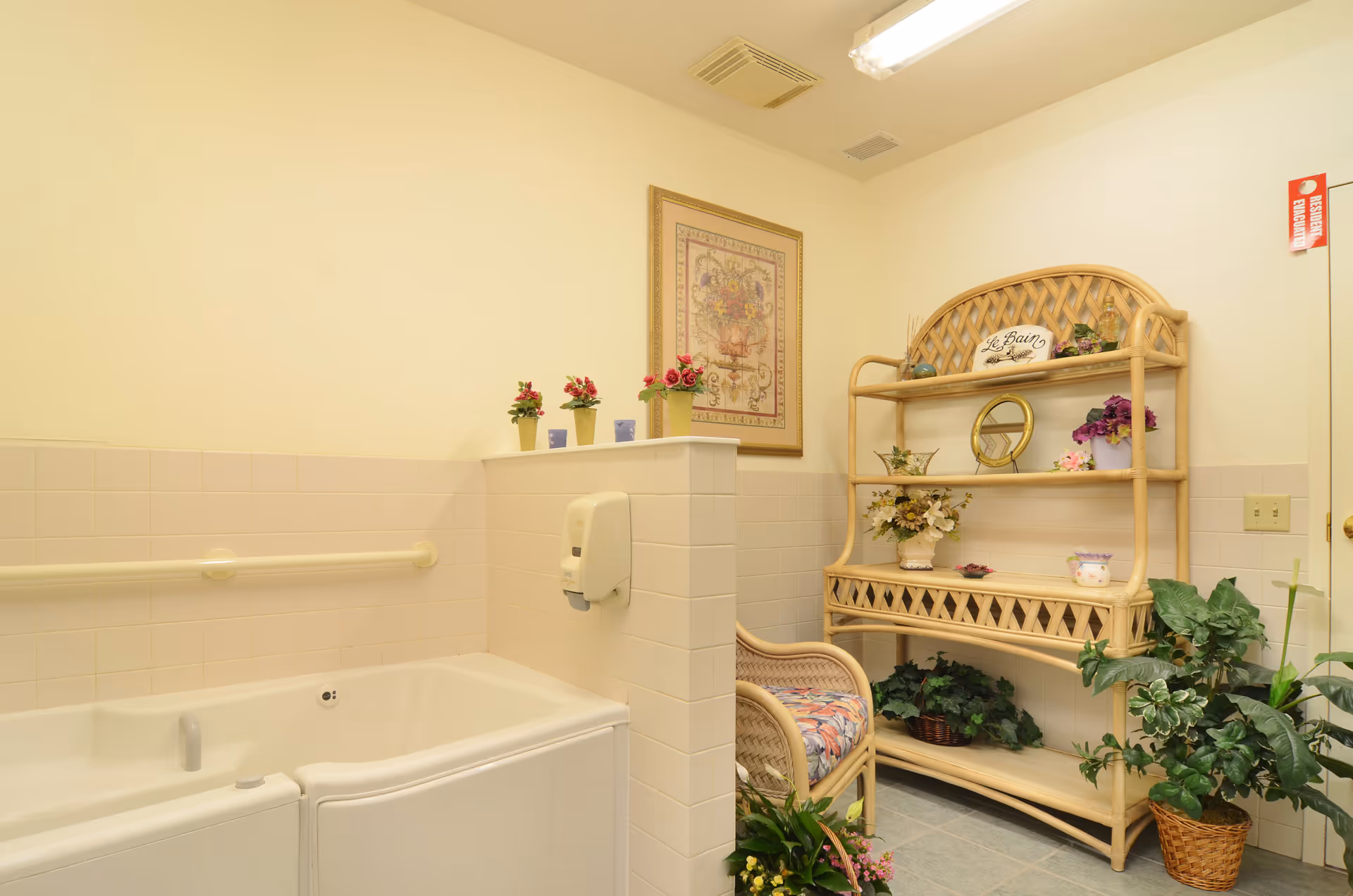 A bathroom area with a walk-in bathtub on the left side, featuring a grab bar and a soap dispenser mounted on a half wall. On the right side, there is a wicker shelving unit decorated with various potted plants, flowers, a small mirror, and a sign that reads 'Le Bain'. A wicker chair with a floral cushion is positioned next to the shelving unit. The walls are light-colored, and there is a framed floral artwork hanging above the half wall.