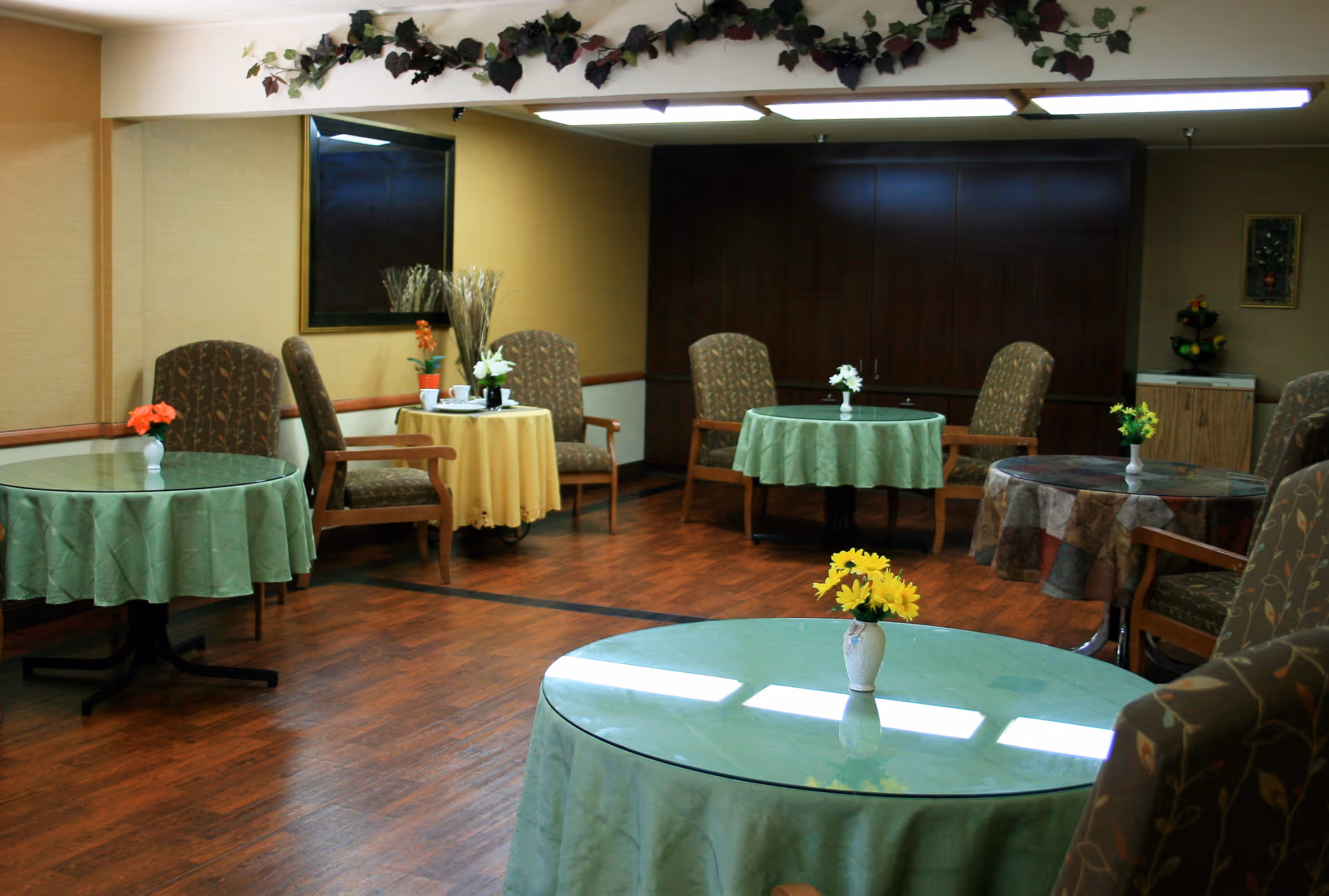 A cozy common area with several round tables covered with green and yellow tablecloths, each adorned with small flower vases. The room has wooden flooring, cushioned chairs with armrests, a large mirror on one wall, and decorative greenery hanging near the ceiling.