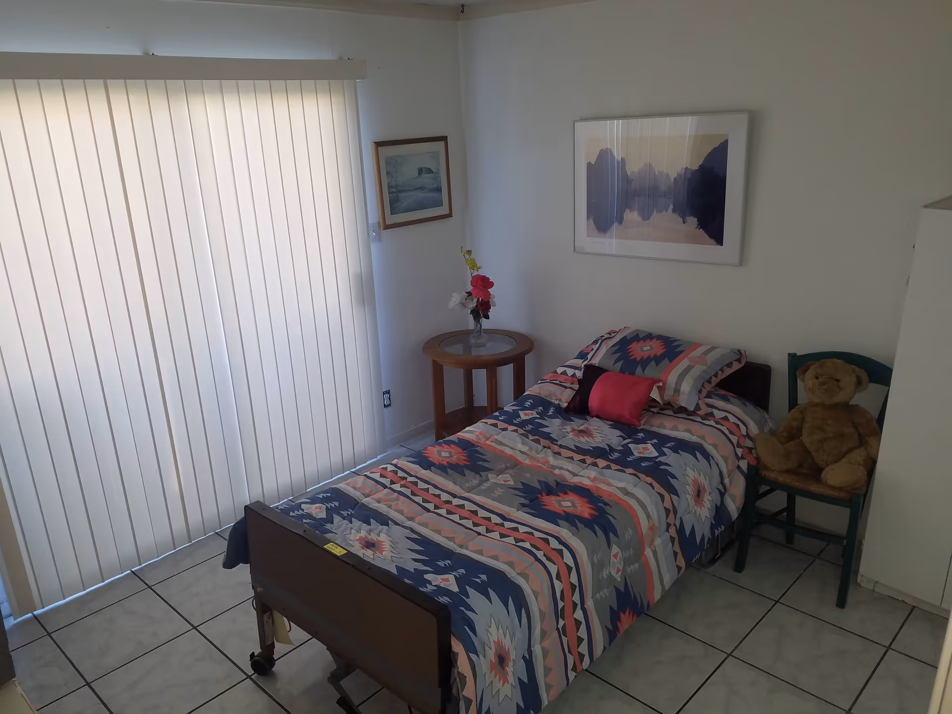 A small bedroom with a single bed covered in a colorful geometric patterned bedspread. There is a small round wooden side table with a vase of flowers next to the bed. A teddy bear sits on a green wooden chair beside the bed. The room has tiled flooring and vertical blinds covering a large window or sliding door. Two framed pictures hang on the white walls.