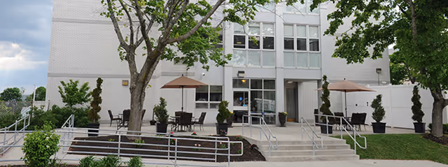 Outdoor patio area in front of a white multi-story building with several tables and chairs under umbrellas, surrounded by trees and greenery, with ramps and stairs leading to the entrance.