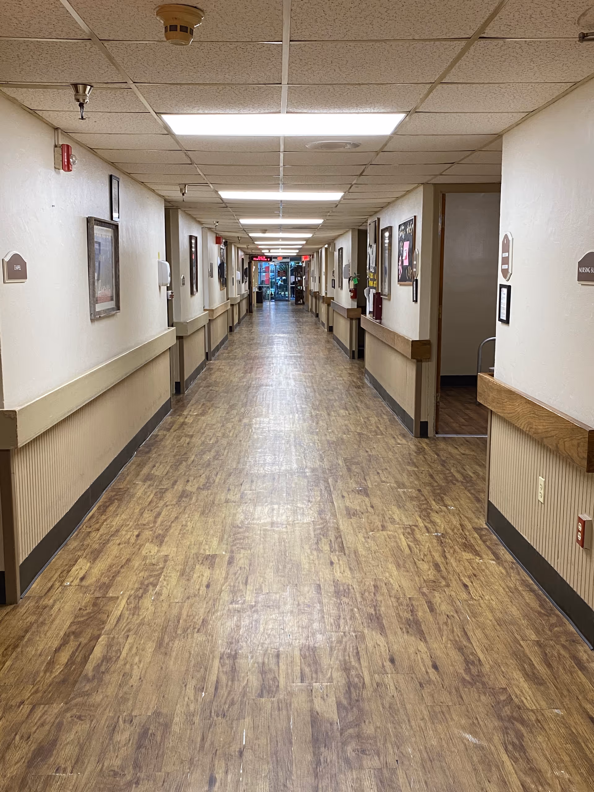 Long well-lit hallway in a senior living facility with wood floors, handrails, and framed artwork along the walls.
