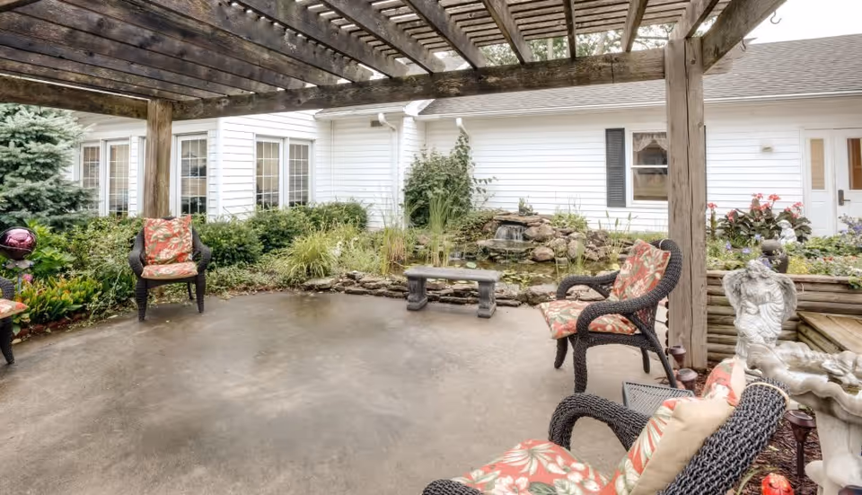 Outdoor patio area with a wooden pergola overhead, several cushioned wicker chairs arranged around the space, a small stone bench, a water feature with a waterfall surrounded by plants, and a white building with windows and a door in the background.