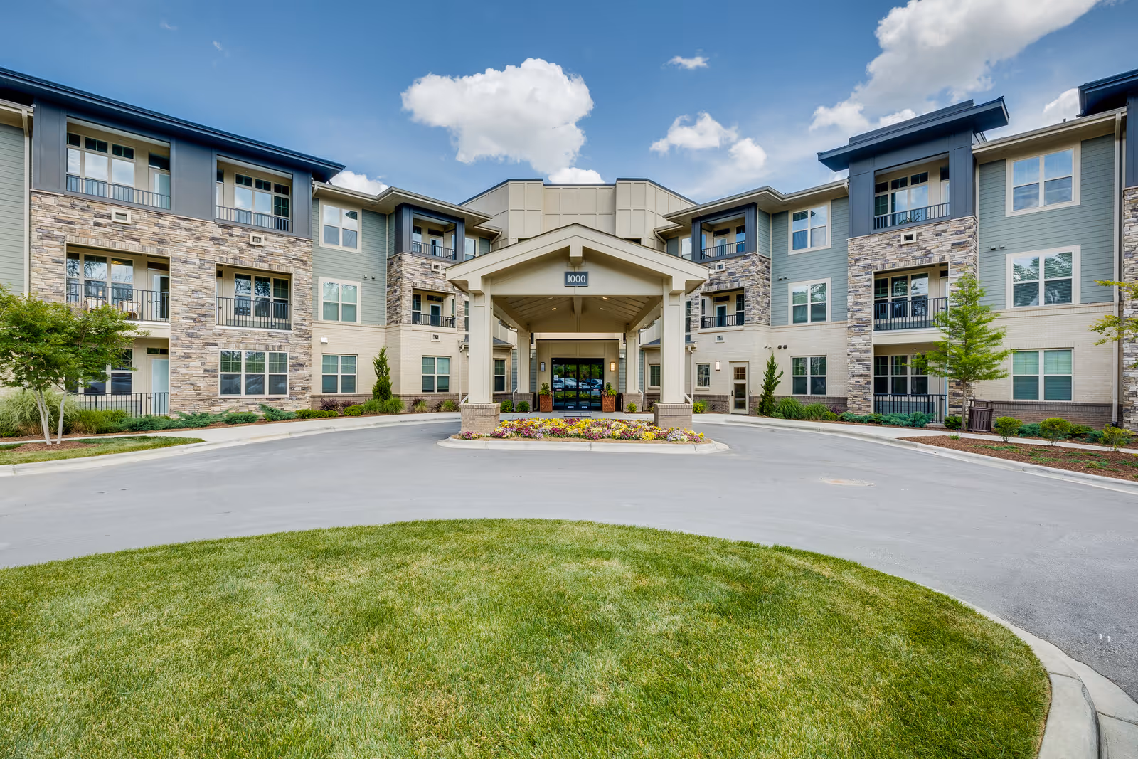 Front exterior view of a modern senior living facility with a circular driveway, landscaped flower beds, and a covered entrance. The building has multiple floors with balconies and a mix of stone and siding on the facade under a partly cloudy sky.