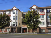 Exterior view of a multi-story residential building with beige and white siding, red trim, and multiple windows. There are two large trees in front of the building, and a street with a sidewalk runs along the front. The building has a sign that reads 'CANNON' above the entrance.