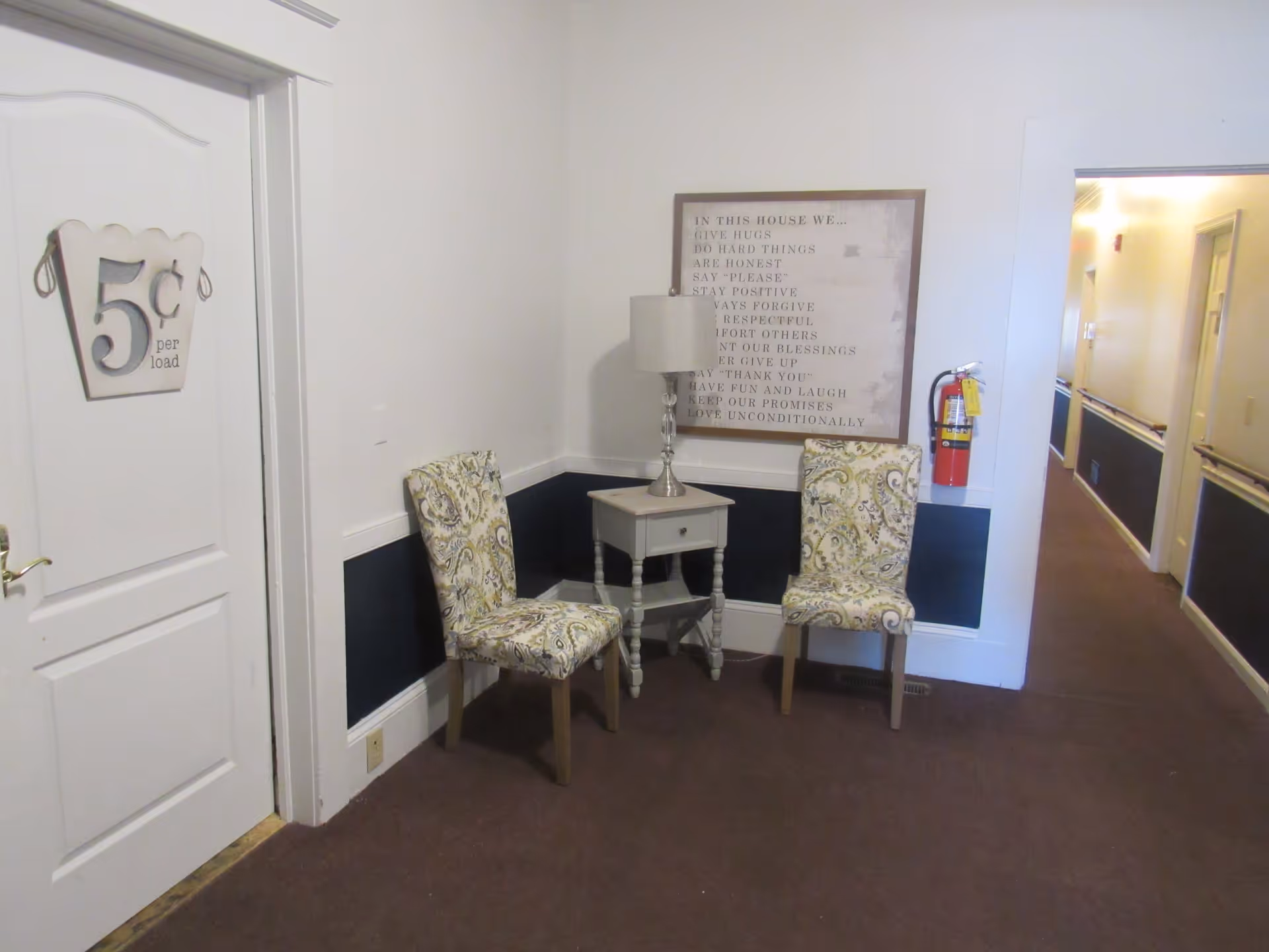 A hallway corner in a senior living facility with two patterned chairs and a small table with a lamp between them. On the wall above the table is a framed sign with inspirational text. A fire extinguisher is mounted on the wall near the entrance to a corridor. A door on the left has a sign that reads '5¢ per load'.