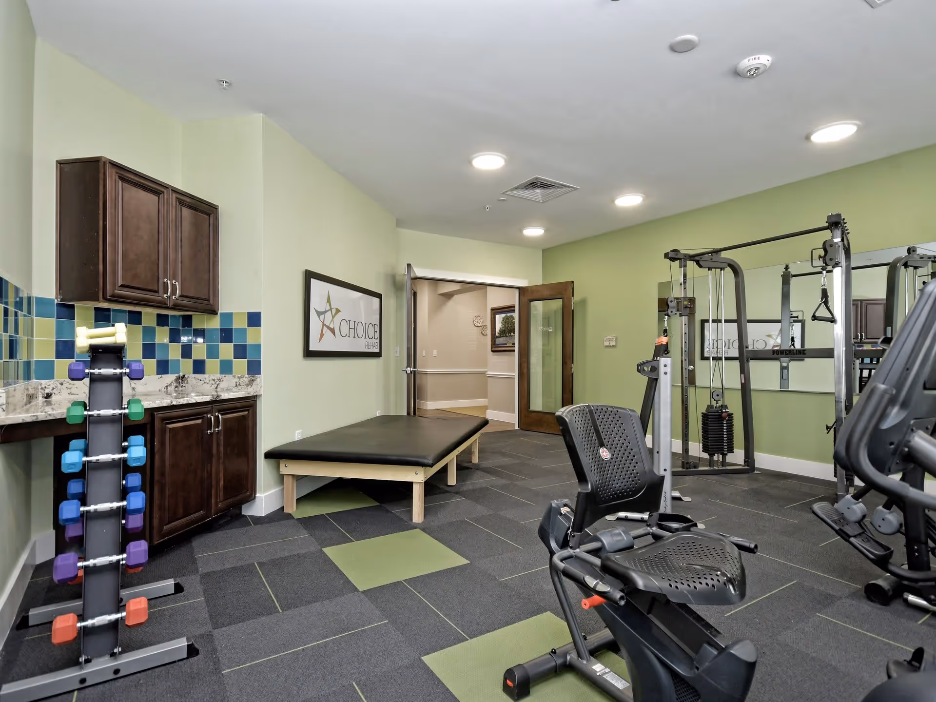 Interior view of a senior living facility fitness room with exercise equipment including a recumbent bike, weight machine, and a rack of colorful dumbbells. The room has green walls, a tiled backsplash above a countertop with cabinets, and a padded exercise table. A framed sign on the wall reads 'CHOICE'.