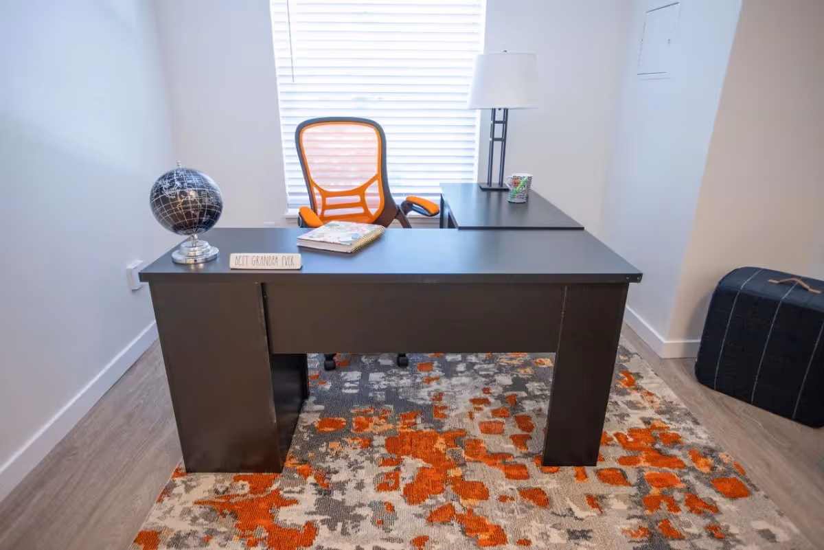 A modern office space with a black desk and an orange mesh office chair. On the desk, there is a black globe, a notebook, a small sign that reads 'BEST GRANDPA EVER', and a table lamp. The room has a window with white blinds and a patterned rug with orange, gray, and beige colors on the floor. A dark blue ottoman is placed in the corner.