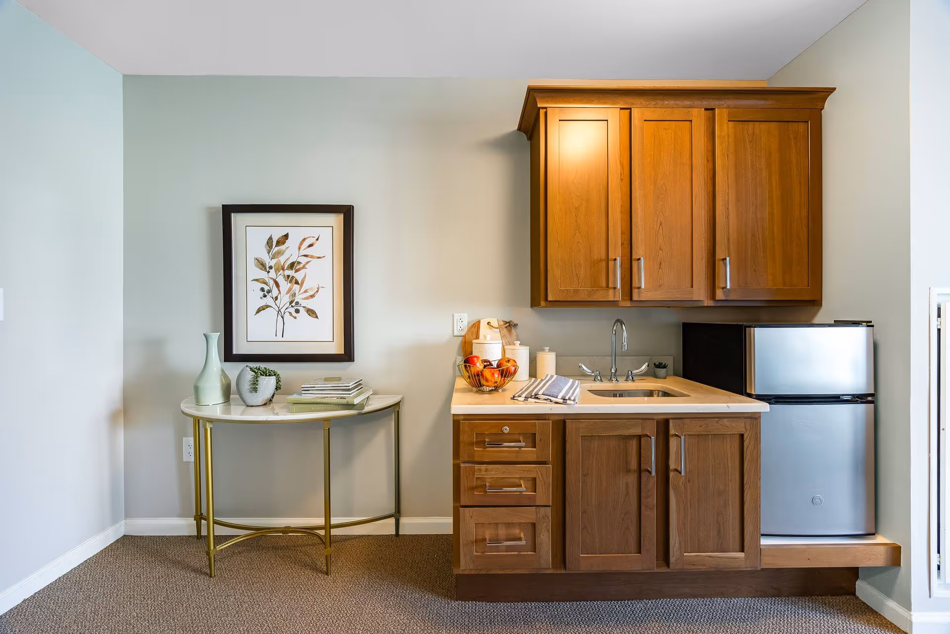 A small kitchenette area with wooden cabinets, a countertop with a sink, a mini refrigerator, and a bowl of apples. To the left, there is a small round table with a vase, a small plant, and some books. A framed botanical artwork hangs on the wall above the table.