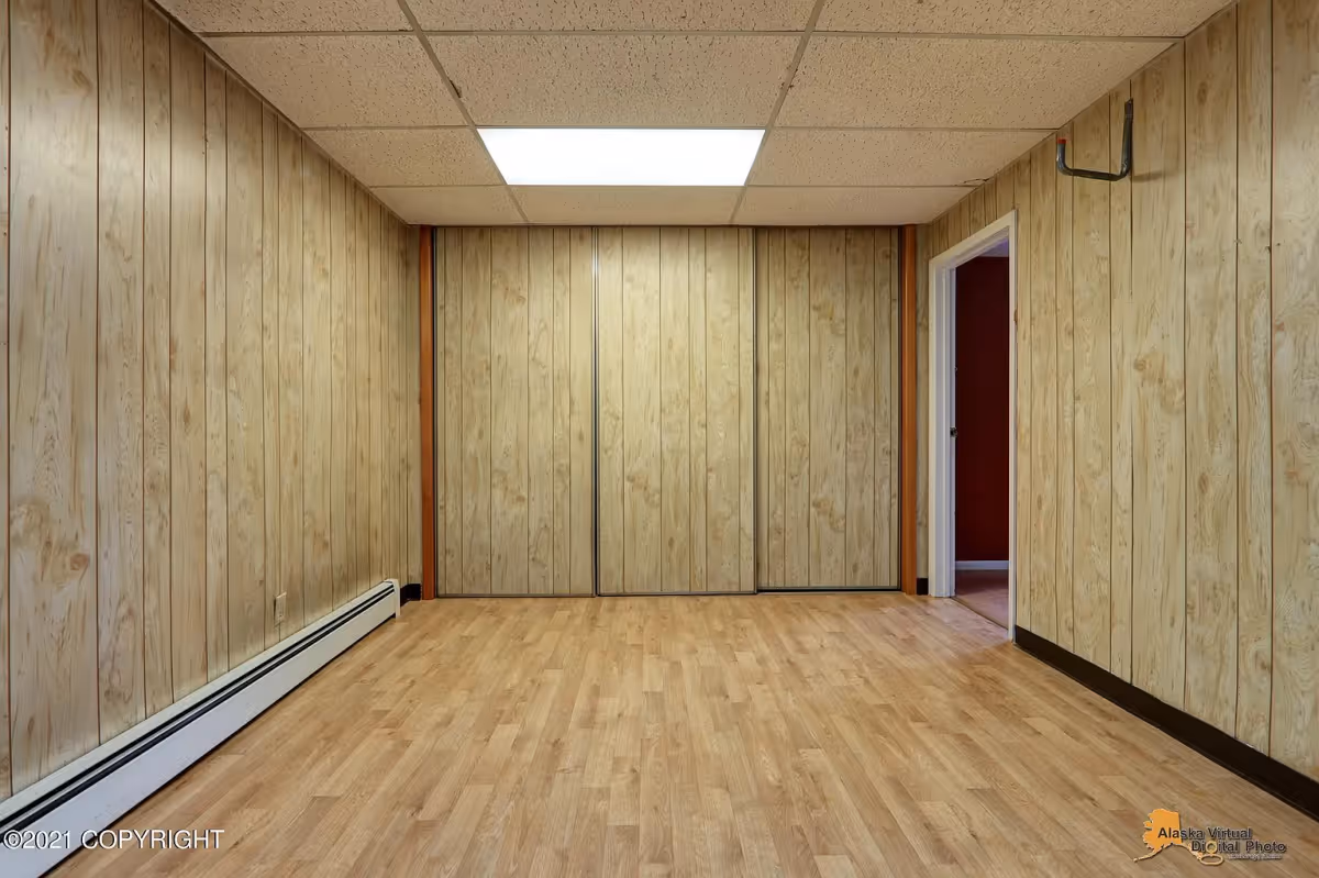 Empty room with wood panel walls, light wood laminate flooring, a fluorescent ceiling light, a baseboard heater along the left wall, a closet with sliding doors on the far wall, and an open doorway leading to another room with red walls.