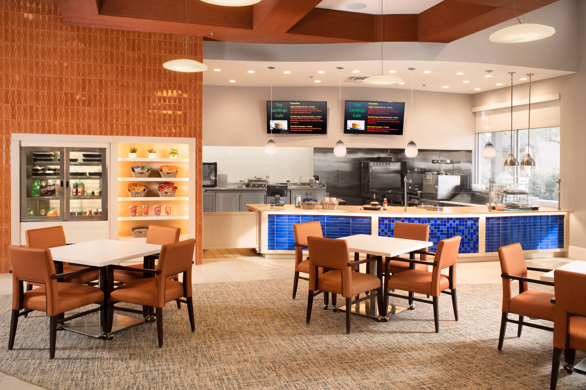 A modern dining area with several square tables and orange upholstered chairs. The background features a serving counter with blue tile accents and a kitchen area behind it. Two digital menu boards are mounted above the counter, and there are pendant lights hanging from the ceiling. A refrigerated display case with snacks and drinks is visible on the left side.