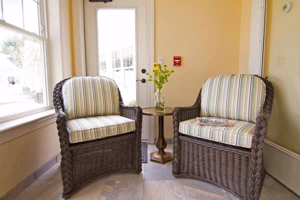 Two wicker armchairs with striped cushions are placed on either side of a small round wooden table holding a vase with yellow flowers. The seating area is next to a window and a glass door, with beige walls and tiled floor.