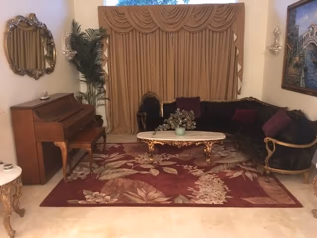 A living room with a dark upholstered sofa and matching armchair, a wooden piano with a bench, a decorative mirror on the wall, a large floral area rug, a marble-top coffee table with a plant centerpiece, heavy draped curtains covering a window, and a framed painting on the wall.