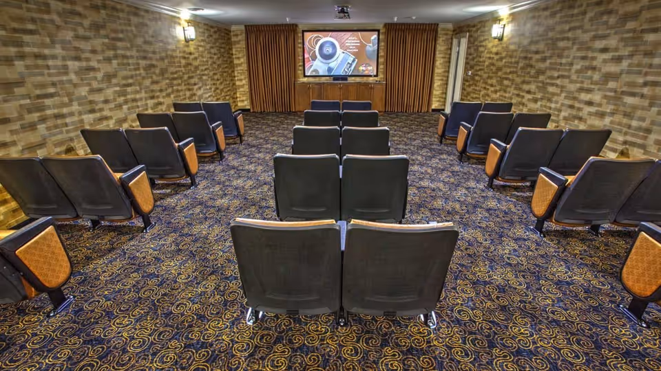 Small screening room with rows of theater seats facing a projector screen and patterned carpet.
