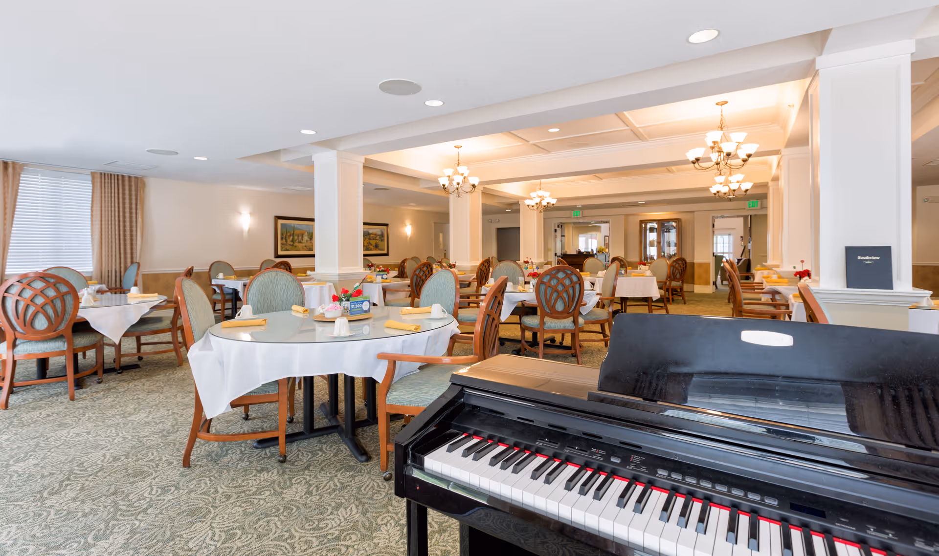A spacious dining room in Southview Assisted Living & Memory Care featuring multiple round tables with white tablecloths and yellow napkins, surrounded by wooden chairs with green upholstery. A black piano is visible in the foreground. The room is well-lit with chandeliers and wall sconces, and decorated with framed artwork and curtains on the windows.