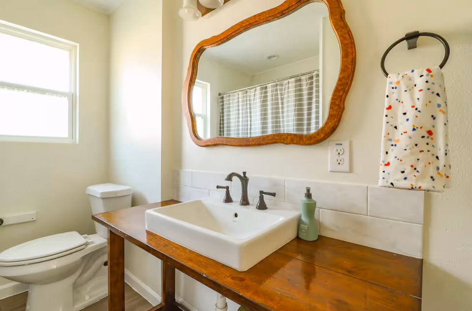 A bathroom with a white toilet next to a wooden vanity with a white rectangular sink. Above the sink is a large, decorative wooden-framed mirror. A towel ring with a white towel featuring colorful specks is mounted on the wall to the right of the sink. A window is visible on the left side, and a shower curtain with a striped pattern is reflected in the mirror.