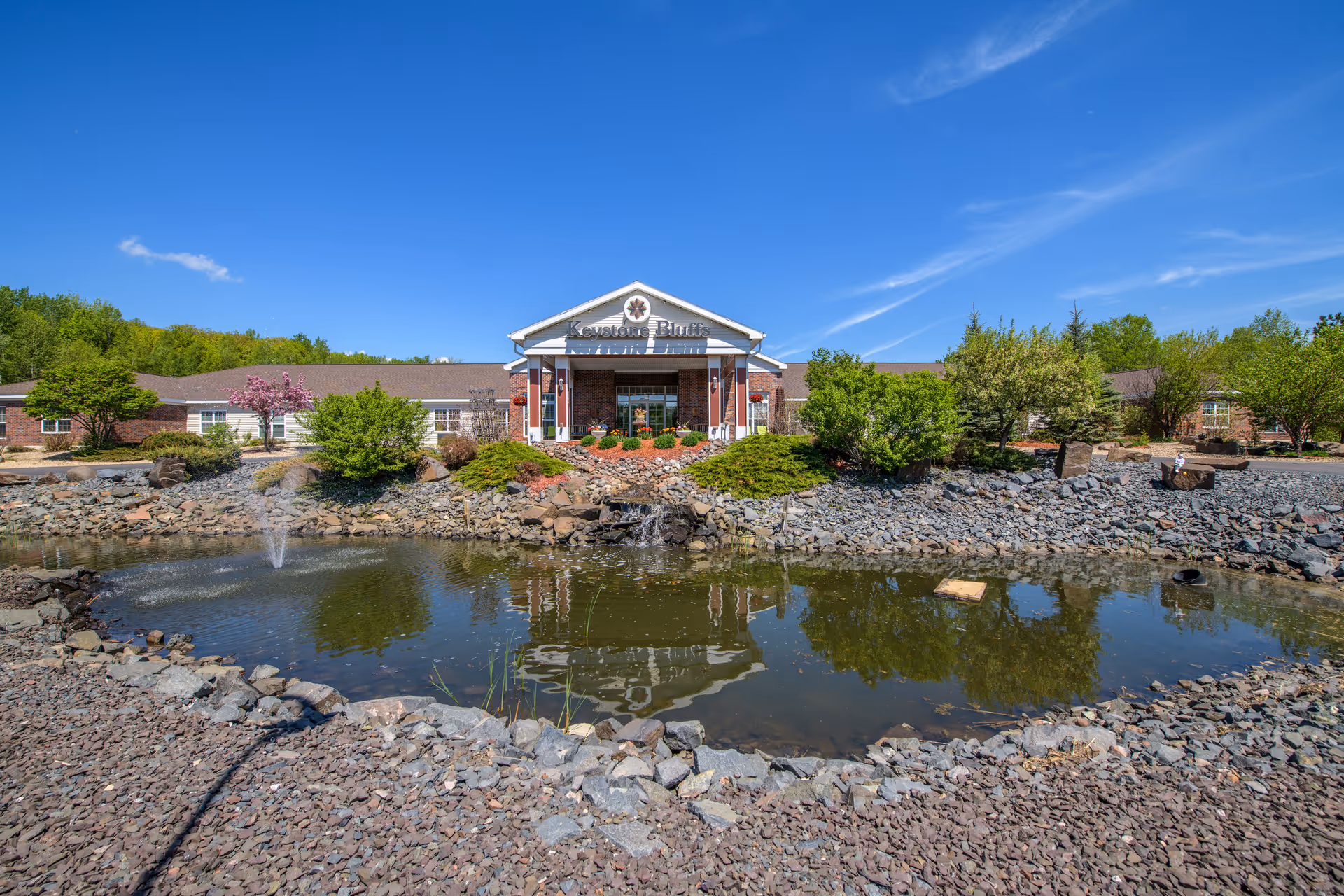 Front exterior view of Keystone Bluffs Assisted Living facility with a pond and fountain in the foreground, surrounded by rocks and greenery under a clear blue sky.