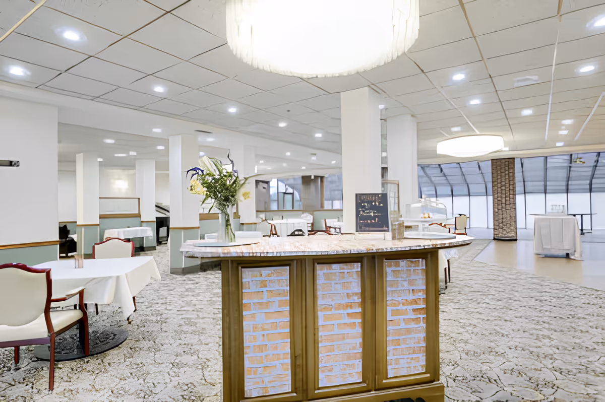 Spacious dining area with tables covered in white tablecloths and chairs with wooden frames and cream cushions. A central island with a marble countertop and brick paneling is decorated with a vase of flowers. The room has a patterned carpet, white walls with green wainscoting, and a ceiling with recessed lighting and large circular light fixtures. Large windows with a curved design are visible in the background.