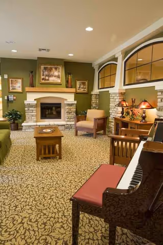 Lounge area with a fireplace, piano, plush chairs, and warm decor in a senior living facility.