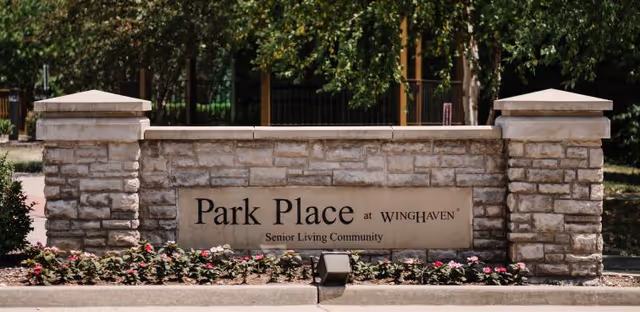 Stone entrance sign for Park Place at Winghaven Senior Living Community surrounded by flowers and greenery.