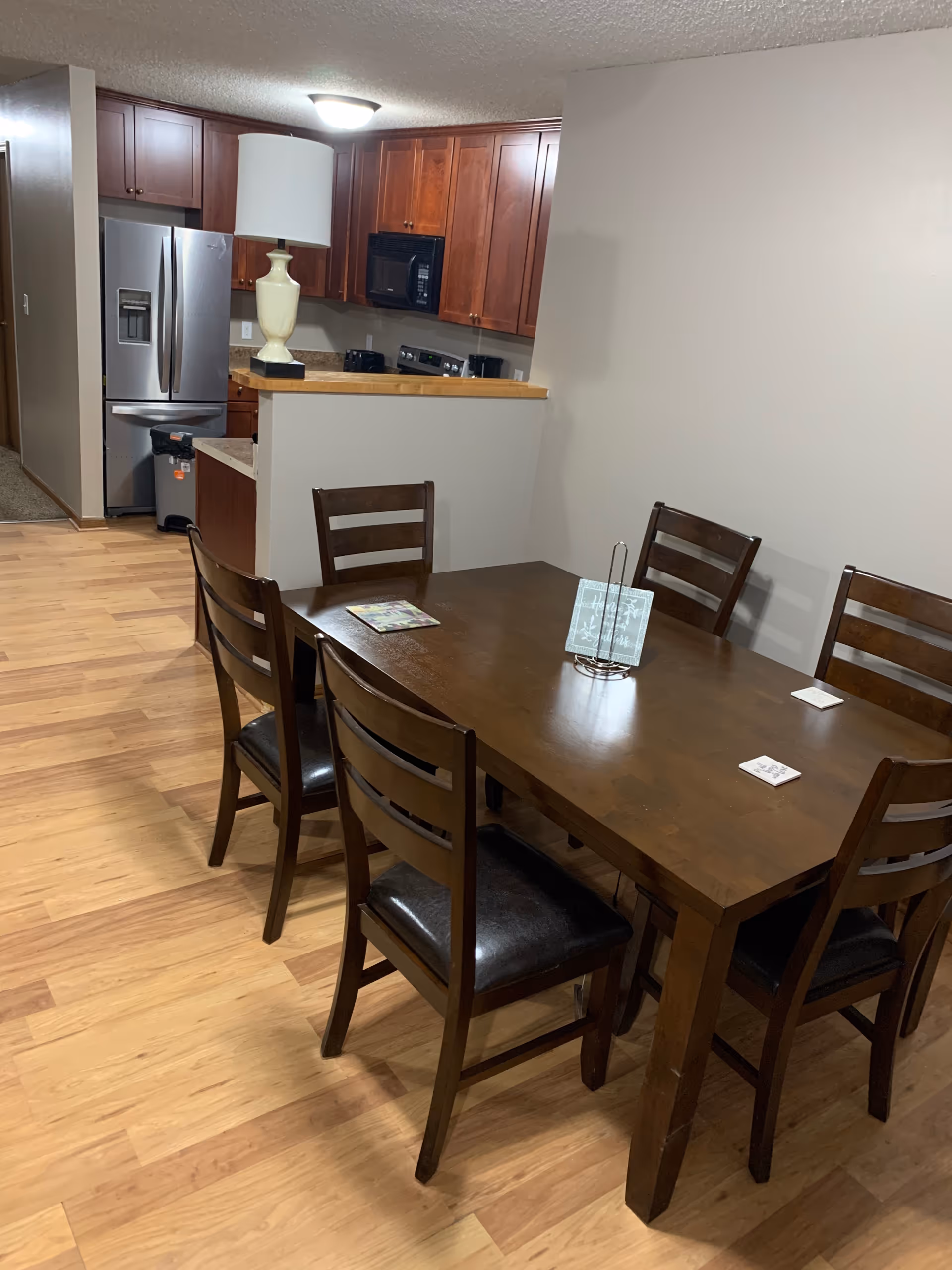 Dining area with a wooden table and six chairs on a wooden floor. In the background, there is a kitchen with wooden cabinets, a stainless steel refrigerator, a microwave, and a stove. A white lamp is placed on the kitchen counter.