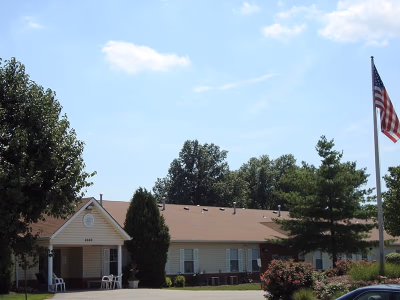 Exterior view of a single-story building with a brown roof and beige siding, surrounded by trees and bushes. There is an American flag on a flagpole to the right and a paved driveway in front. The sky is clear with some clouds.