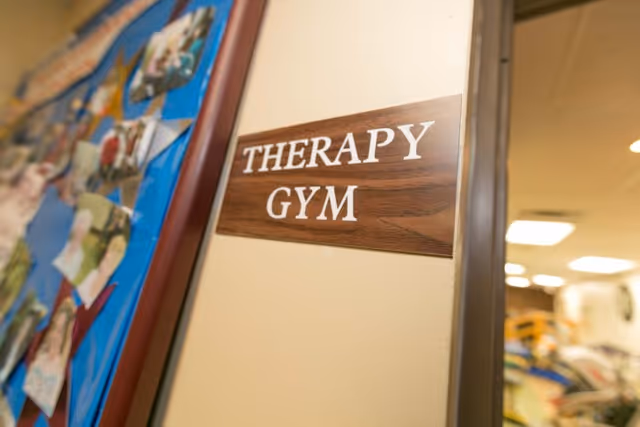 A close-up view of a wooden sign on a beige wall that reads 'THERAPY GYM'. To the left, there is a bulletin board with various photos pinned on it. The background shows a blurred view of gym equipment and ceiling lights inside the therapy gym.