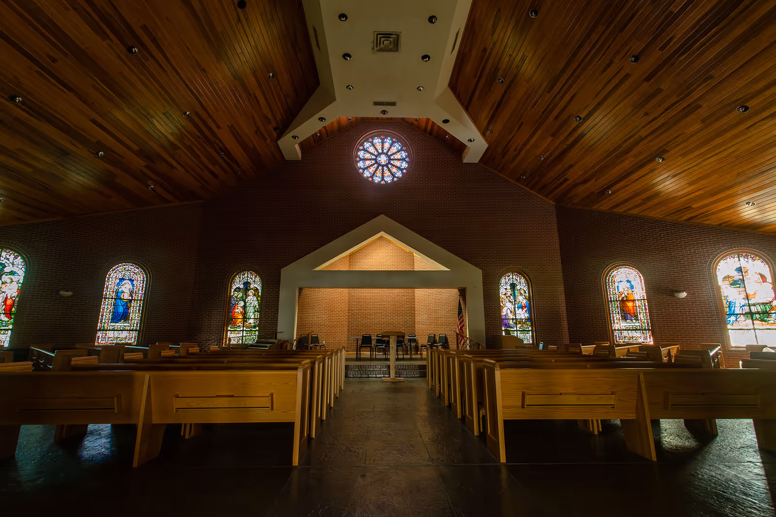 Interior view of a chapel with wooden pews, stained-glass windows along the walls, and a small front stage.