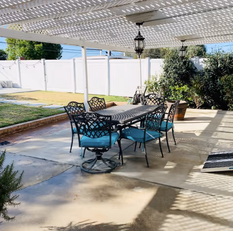 Outdoor patio area with a metal table and six matching chairs with blue cushions under a white pergola. The patio is surrounded by a white fence and some greenery, including bushes and a tree.