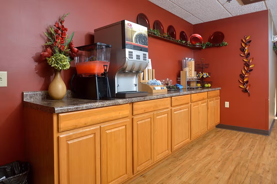 A beverage and snack station with a wooden cabinet base and granite countertop against a red wall. The station includes a drink dispenser with a red beverage, a coffee machine, cereal dispensers, cups, and some decorative items including a vase with flowers and a wall-mounted metal leaf decoration.